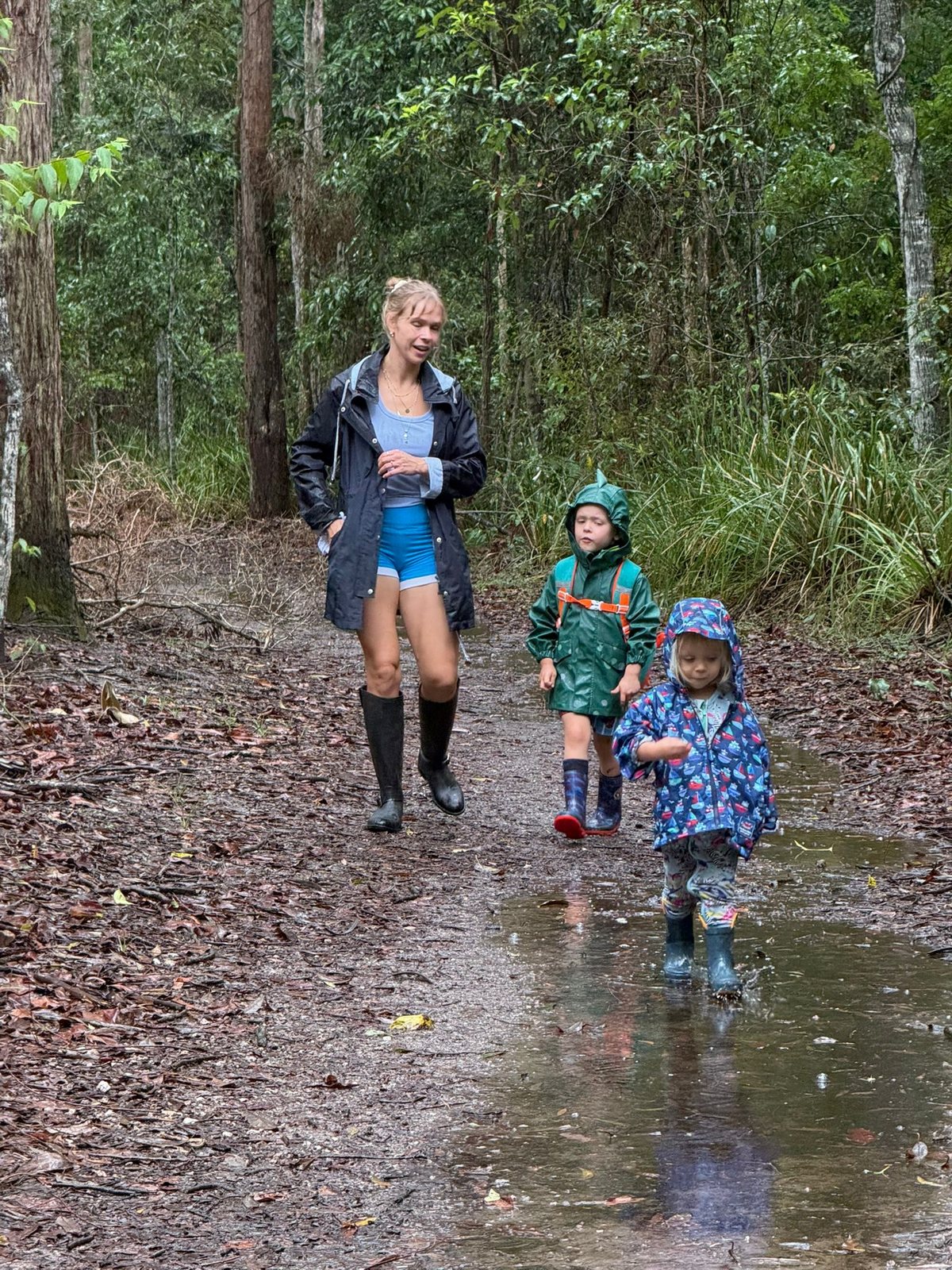 Sally walking through a rainy forest with her two boys in gumboots and raincoats, splashing through puddles